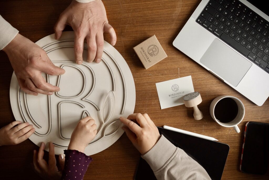 Overhead shot of multiple hands assembling a wooden 'B' logo puzzle on a desk, surrounded by branding materials, a laptop, and coffee, symbolizing collaborative brand development.