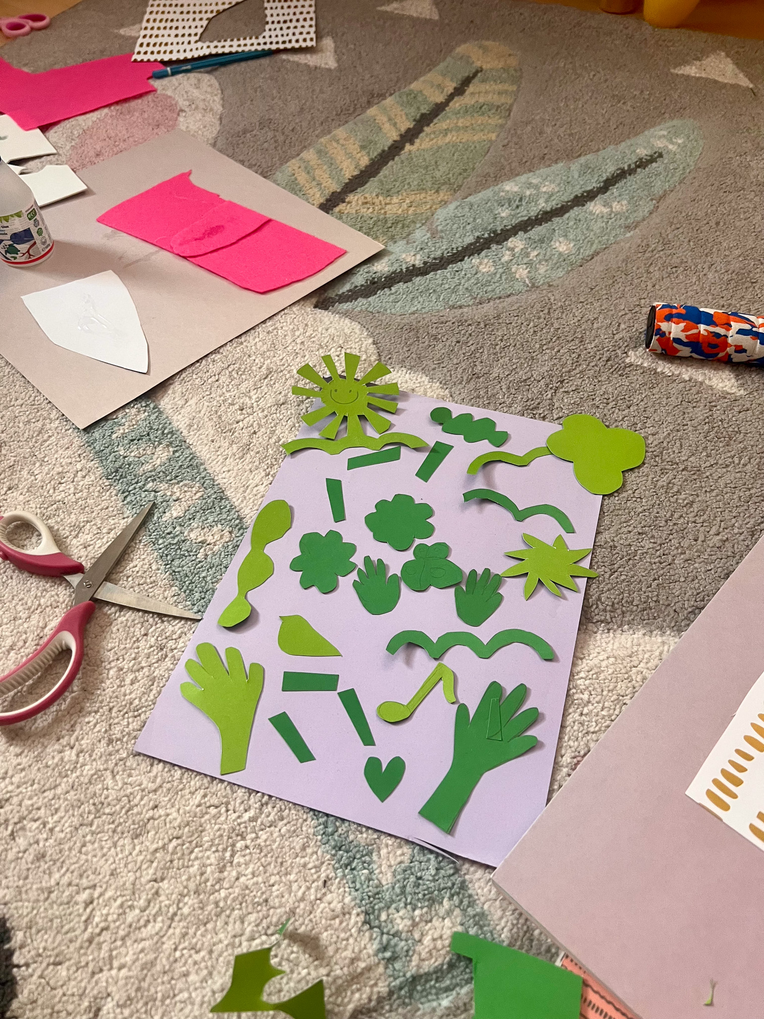 Colorful craft materials including green paper cutouts arranged on a purple sheet, scissors, and glue on a patterned rug.