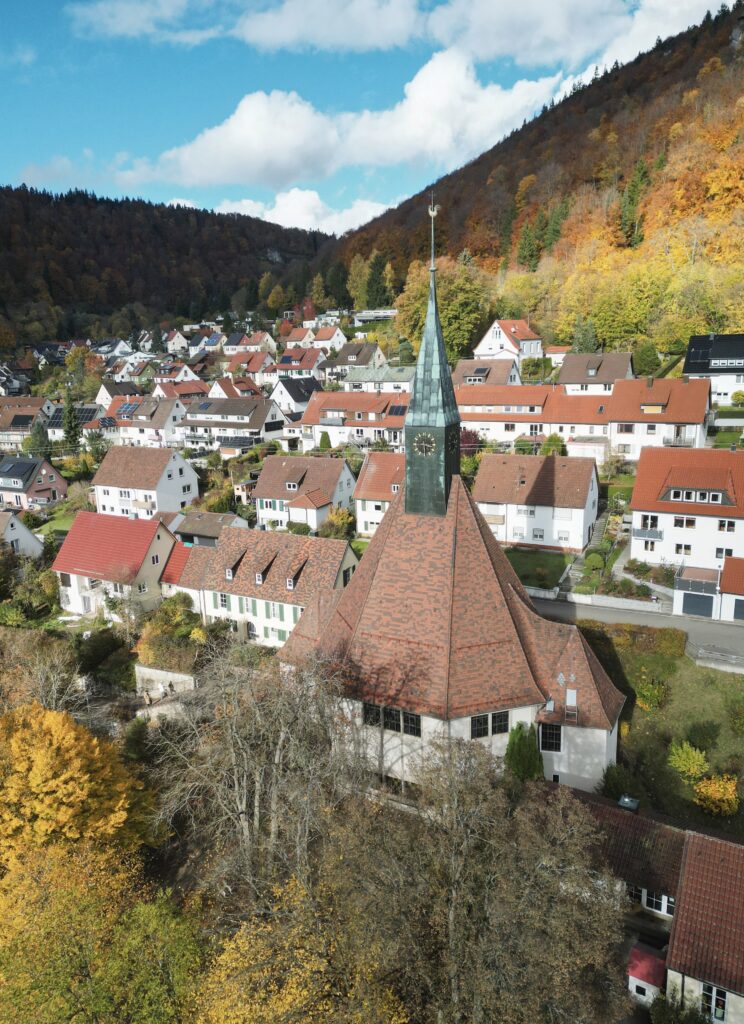 Aerial view of a traditional German village with a prominent church spire nestled in a valley surrounded by vibrant autumn forest foliage.