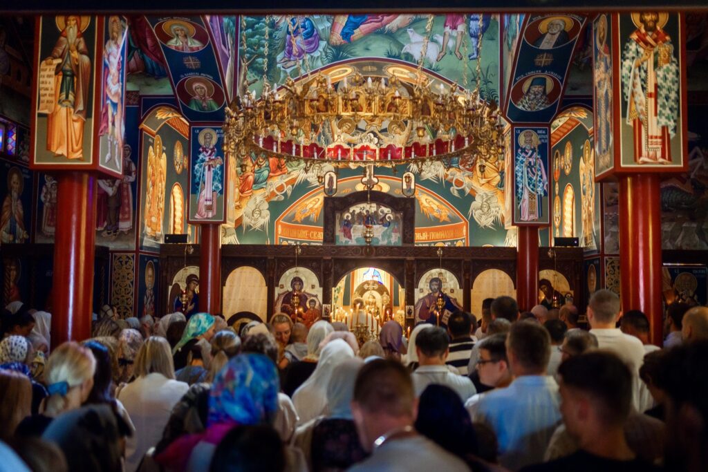 A crowded church interior featuring vibrant frescoes and religious icons, with worshippers engaged in prayer.