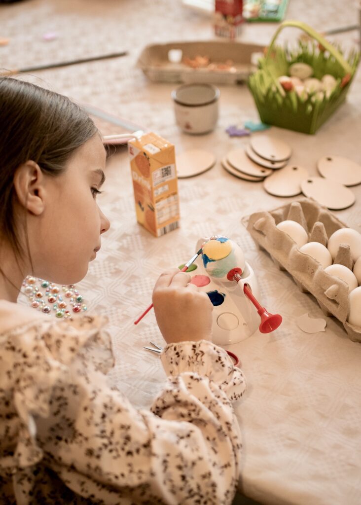 A young girl focused on painting a decorative Easter egg with a small brush at a craft table filled with art supplies and egg cartons.