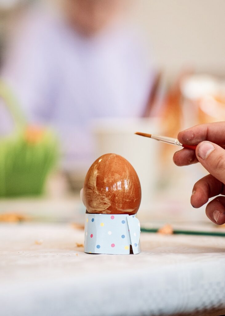 A person carefully painting a brown Easter egg with a fine brush, set in a decorative polka-dot holder during a creative holiday craft session.