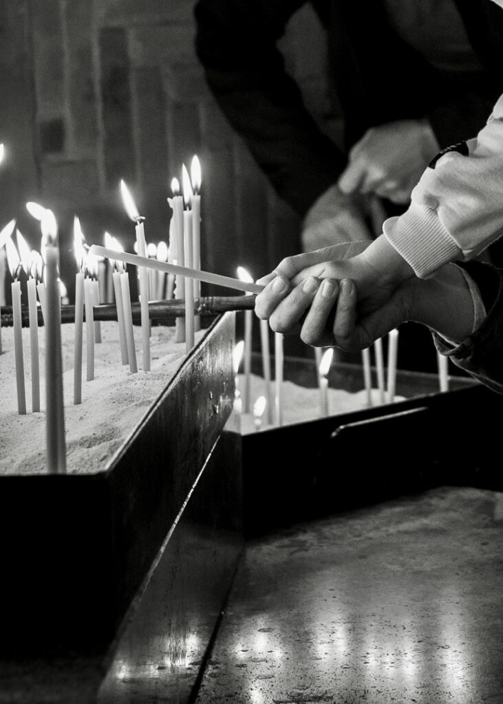 A close-up black and white shot of hands lighting thin prayer candles in a church, symbolizing faith, tradition, and spiritual reflection.