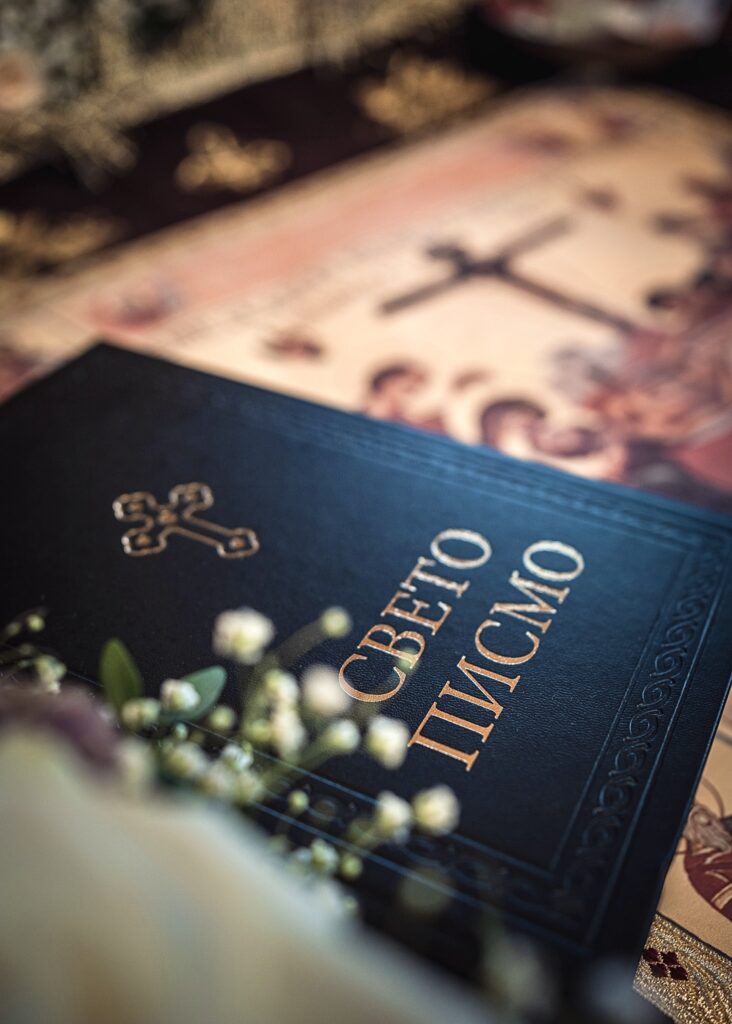 A black leather-bound Holy Bible with gold lettering and a cross symbol, resting on a church altar with soft focus floral arrangements nearby.