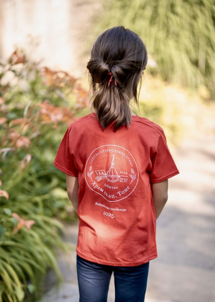 A young girl wearing a red t-shirt with a custom church logo design, standing in a garden setting for a professional community event portrait.