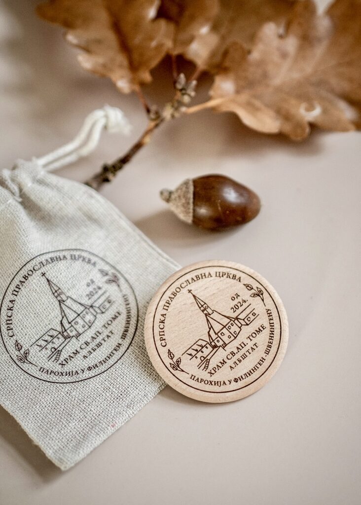 A wooden commemorative coin and a linen pouch featuring the Serbian Orthodox Church logo, displayed with an oak leaf and acorn on a neutral surface.