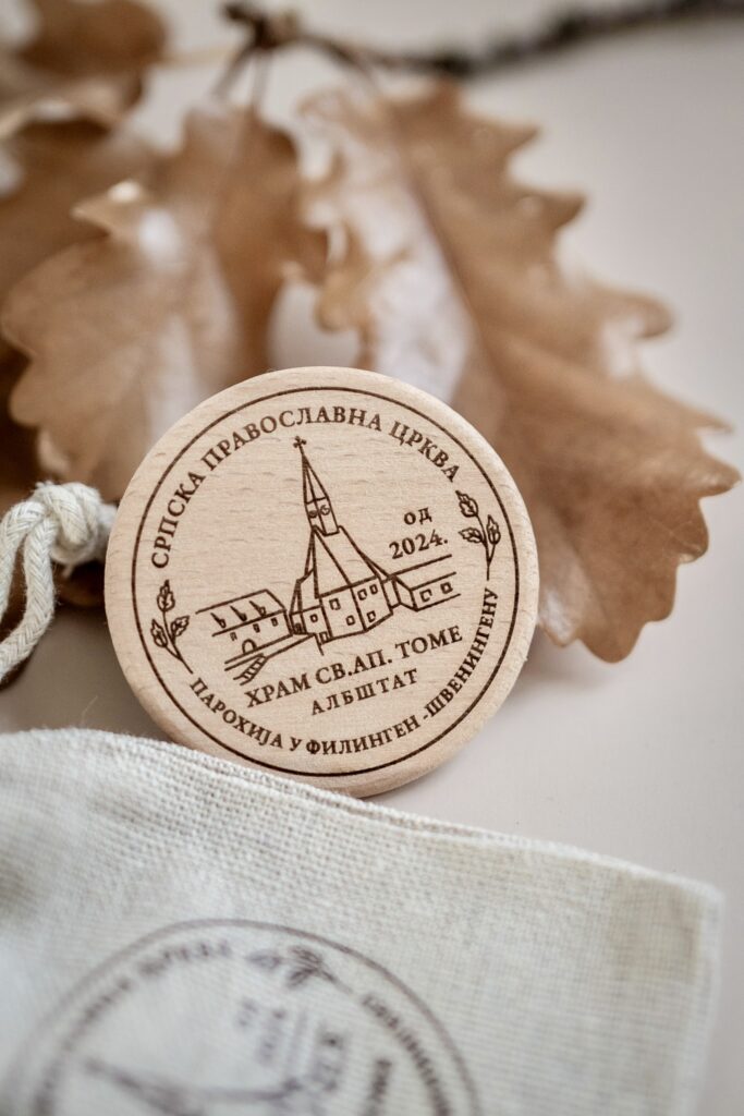 A decorative wooden coin featuring an engraved Serbian Orthodox church design, displayed with dried oak leaves and a small linen fabric pouch.
