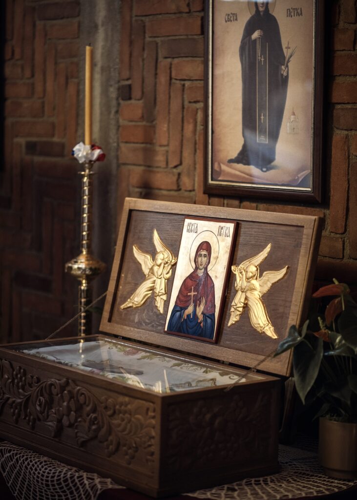 An ornate wooden reliquary featuring a religious icon of Saint Petka, displayed in a church with a candle and a framed portrait on a brick wall.