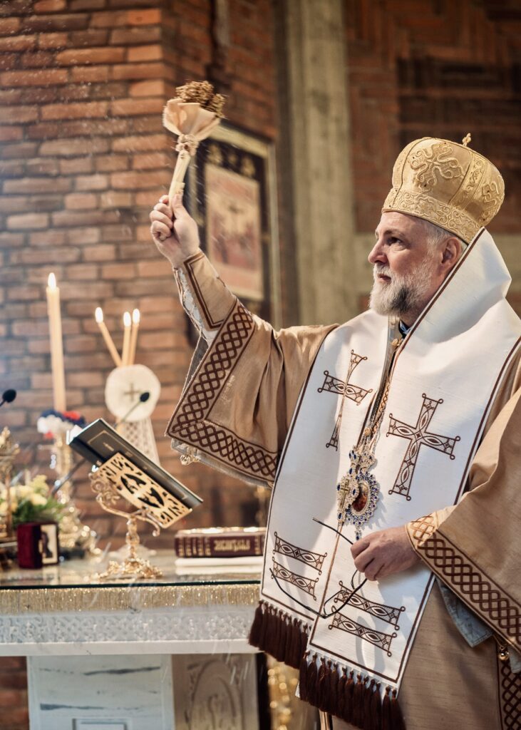 An Orthodox bishop in traditional vestments performing a holy water blessing with an aspergillum during a solemn church liturgy service.