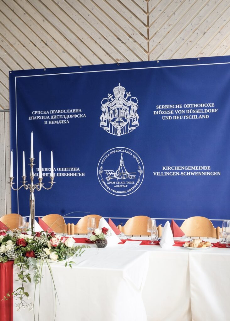 A formal banquet table set for a Serbian Orthodox Church event in Villingen-Schwenningen with a blue branded backdrop and floral centerpiece.