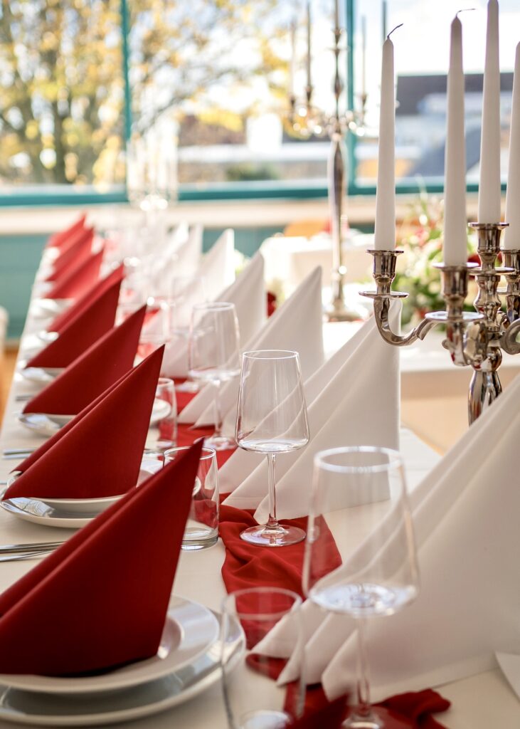 Elegant dining table set for a formal event with red napkins, white linens, crystal glassware, and a silver candelabra in a bright venue.
