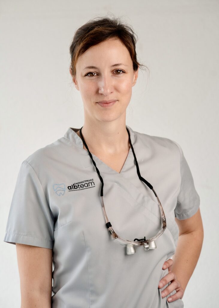 Professional dental practitioner wearing scrubs and magnifying glasses, smiling confidently in a clinical setting.