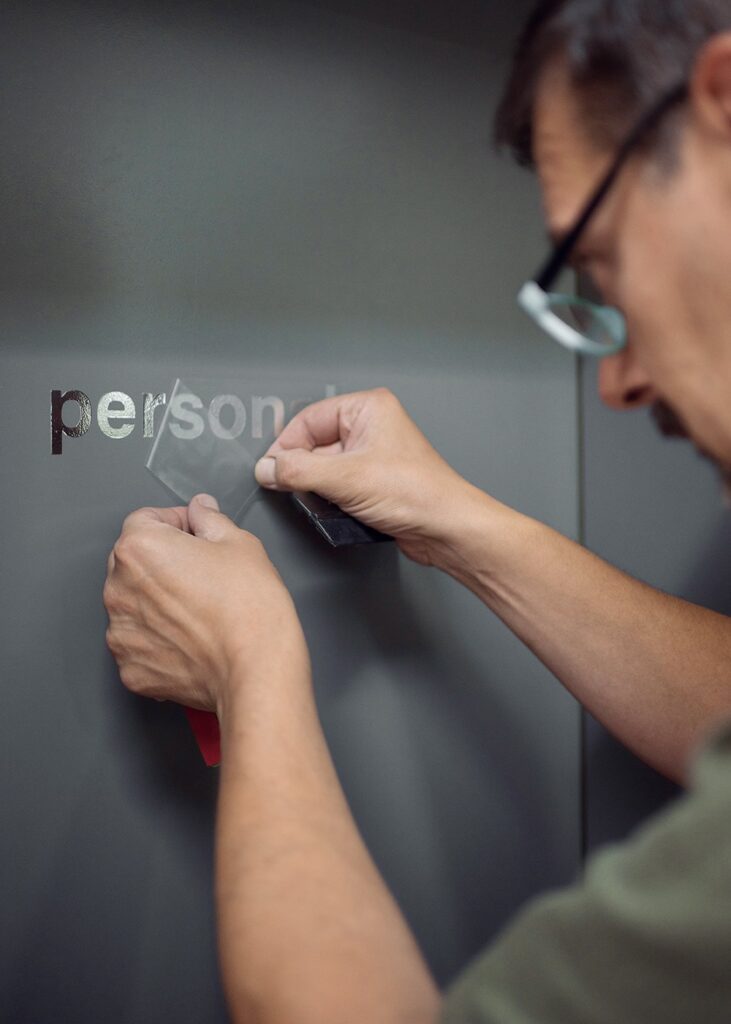 A professional technician carefully applies custom vinyl lettering to a smooth grey surface using a squeegee tool for precise branding.