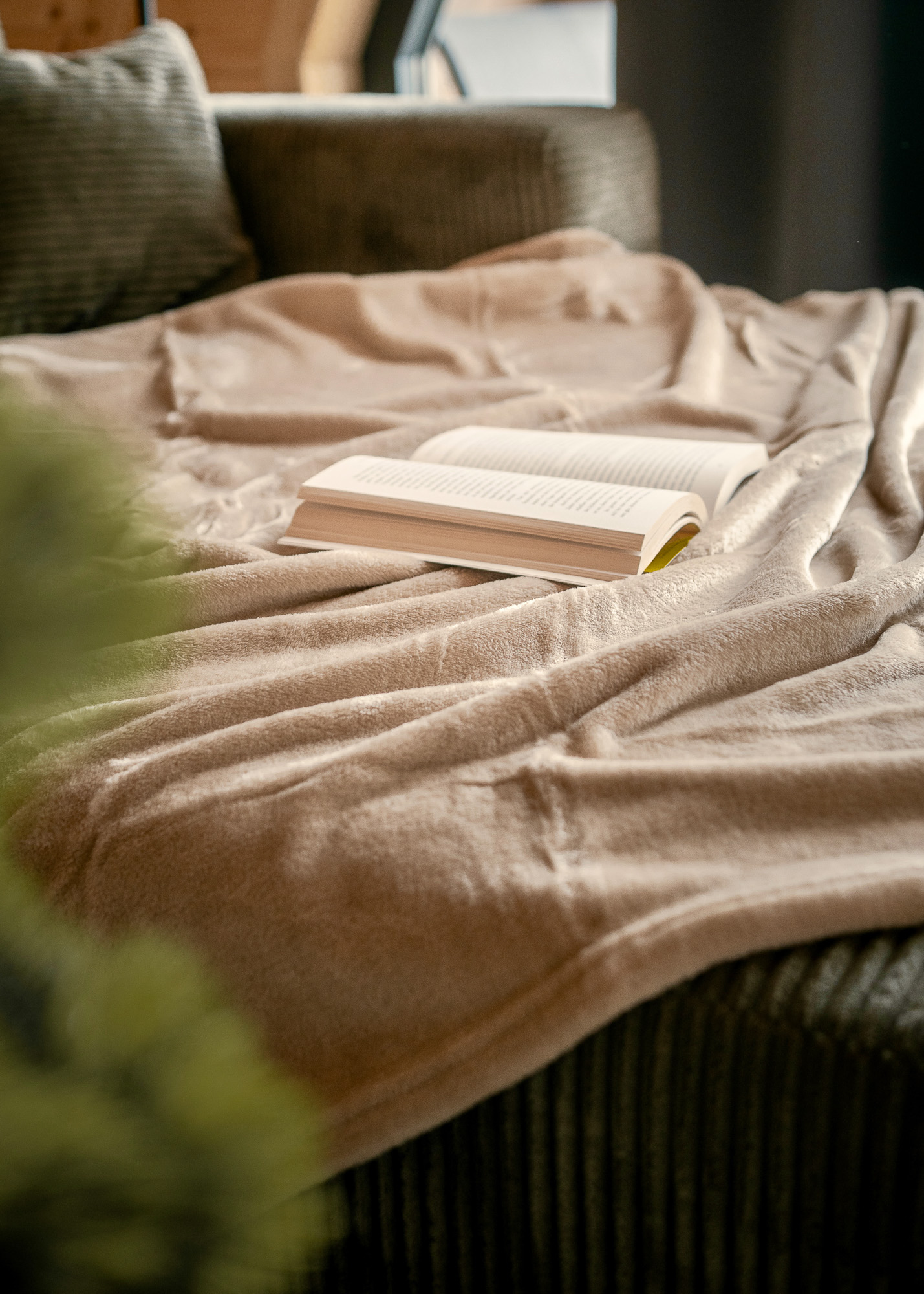 An open book rests on a soft, beige fleece blanket draped over a dark green sofa in a cozy, sunlit living room setting with a blurred foreground.