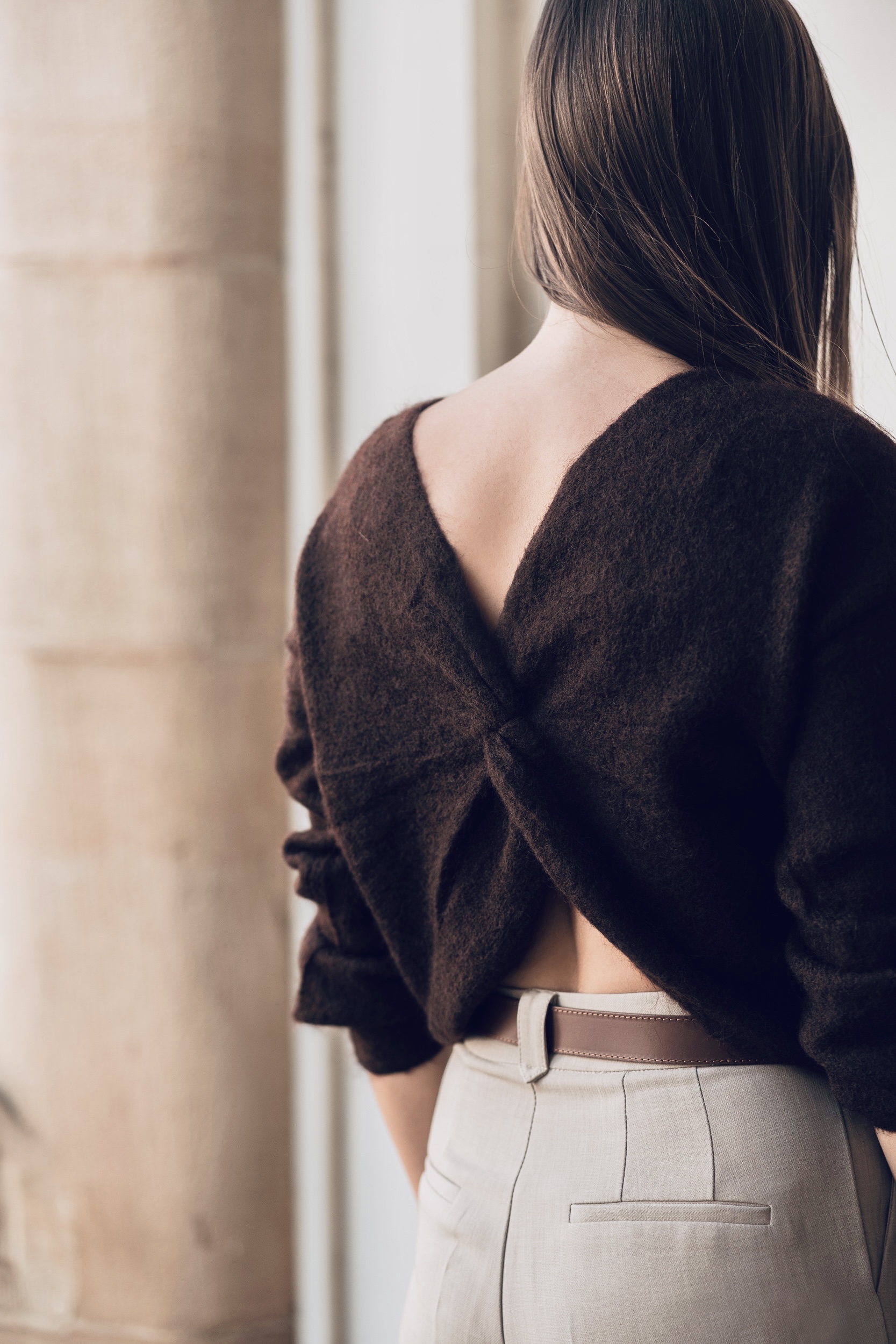 A woman seen from behind wearing a dark brown, long-sleeved sweater with a twisted open-back design, paired with light-colored tailored trousers.
