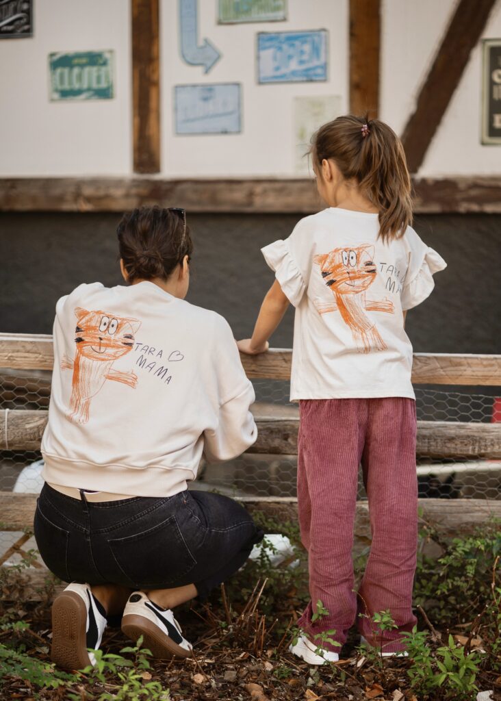 A woman and a young girl wearing matching shirts with a hand-drawn orange cat illustration, standing together by a wooden fence at an outdoor area.