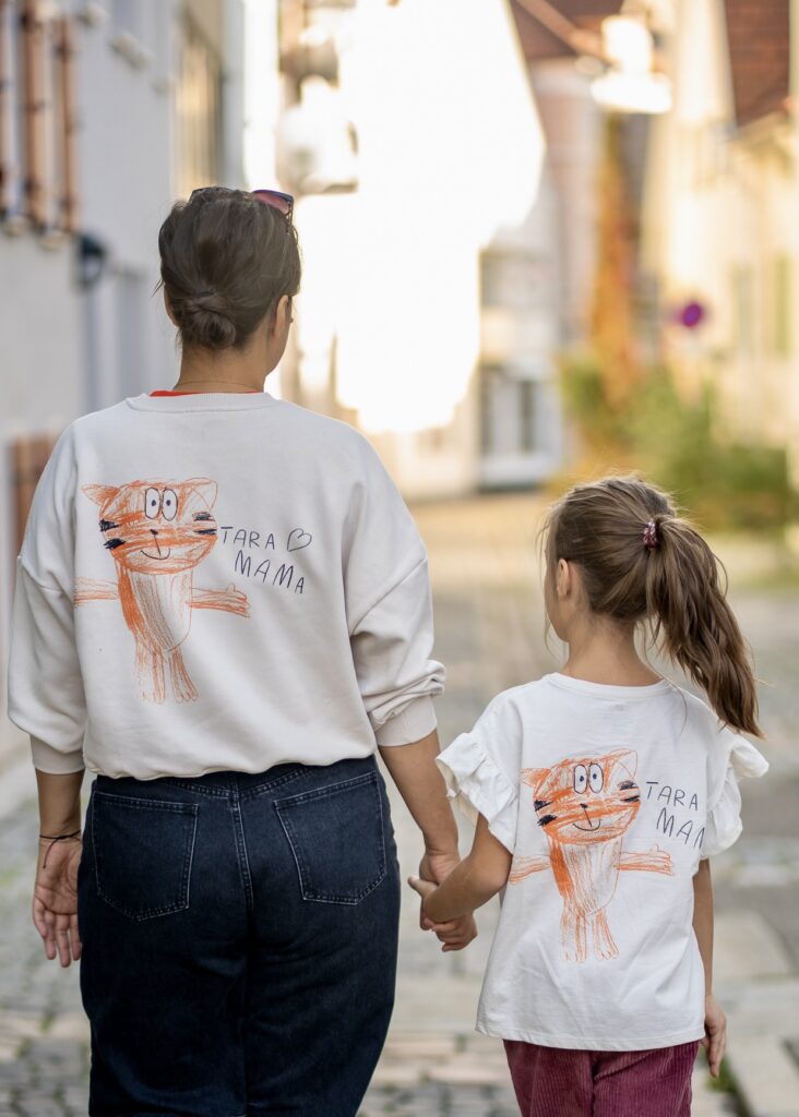 A woman and a young girl walk hand-in-hand, both wearing matching white shirts featuring a child's drawing of a cat and the text Tara Mama.