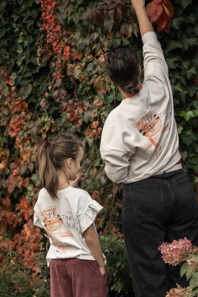 A woman and a young girl wearing matching cream-colored shirts with a hand-drawn cat illustration, standing before a lush wall of autumn foliage.