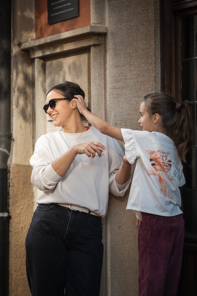 A woman in sunglasses smiles while a girl playfully tugs at her hair, both enjoying a sunny day outdoors.