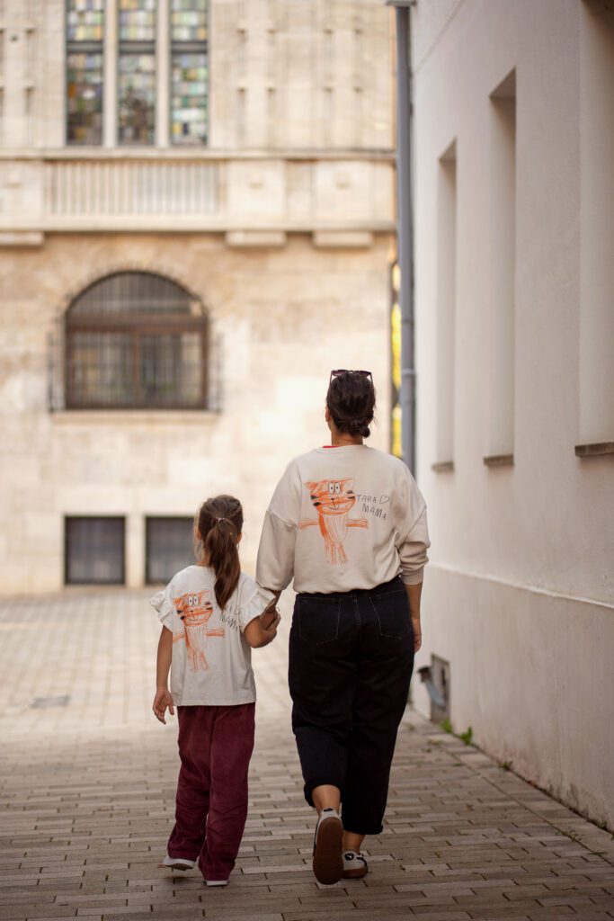 A mother and daughter walk hand-in-hand down a cobblestone path, both wearing matching white shirts featuring a hand-drawn orange cat illustration.
