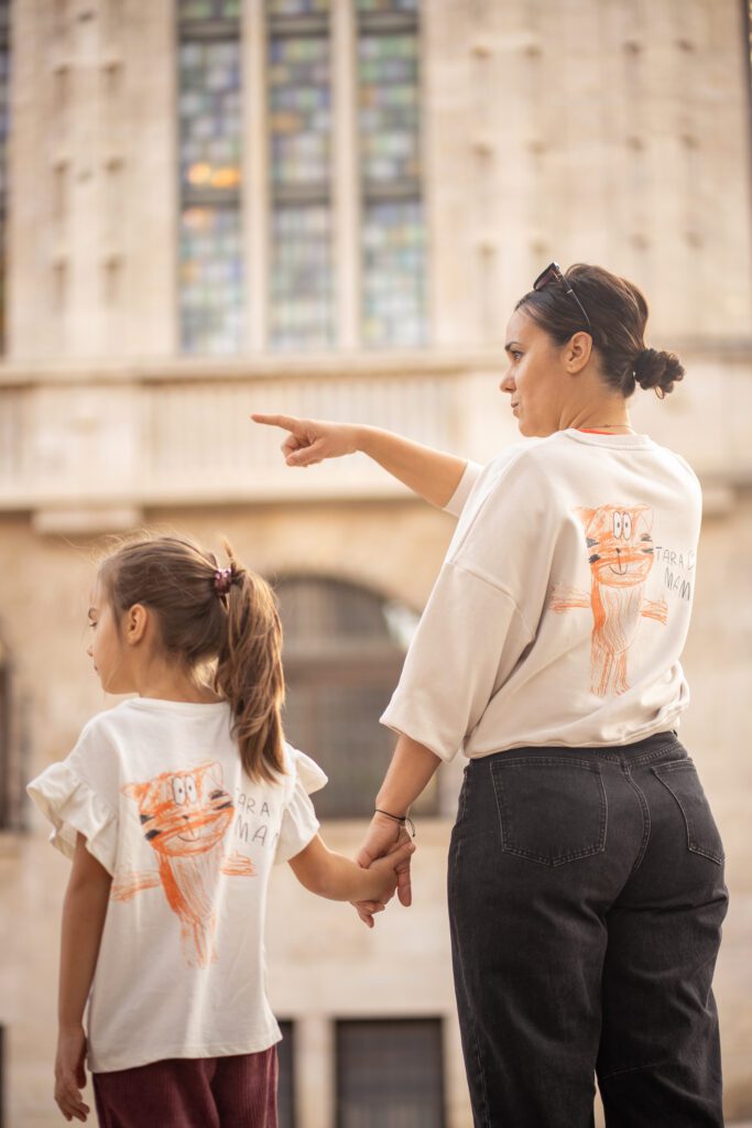 A woman and a young girl holding hands and wearing matching shirts with cat drawings, standing in front of a historic building as she points away.