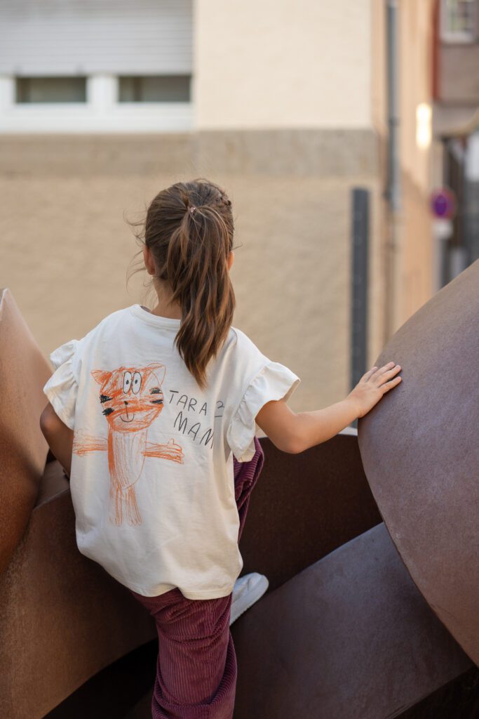 A young girl with a ponytail, wearing a white t-shirt with a hand-drawn orange tiger, stands outdoors leaning against a large, curved metal sculpture.