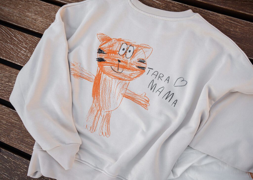 A light beige sweatshirt featuring a child's orange crayon drawing of a cat and the handwritten text TARA MAMA with a heart on a wooden surface.