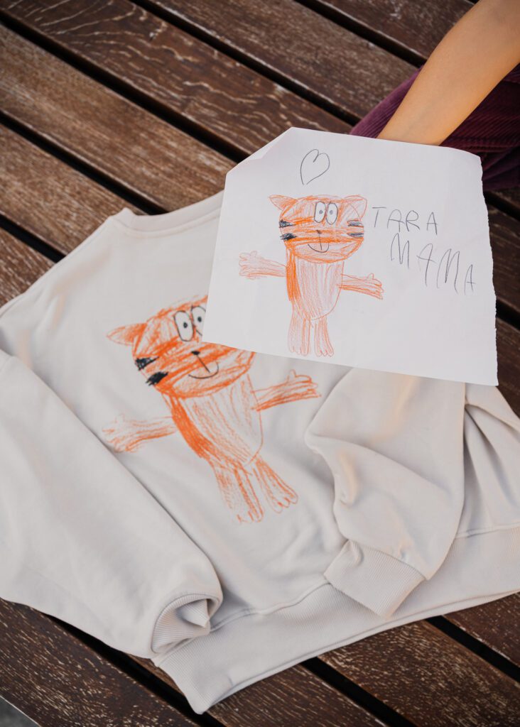 A hand holds a drawing of an orange tiger above a beige sweatshirt that features the exact same child's drawing printed on its fabric surface.