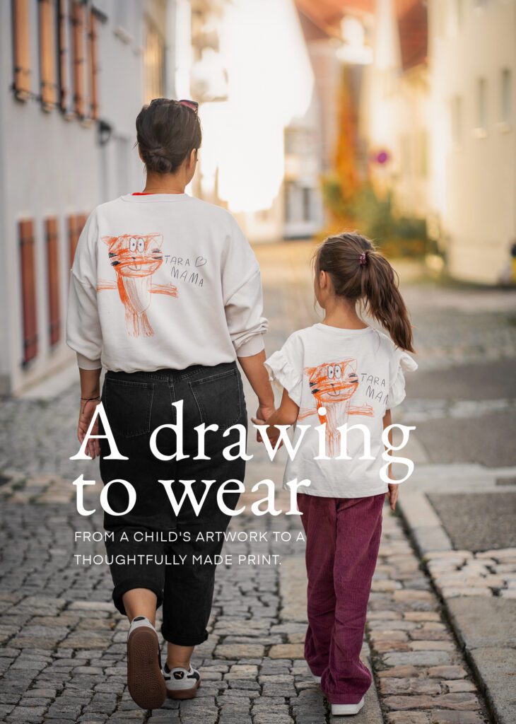 A mother and daughter walk hand-in-hand on a cobblestone street, wearing matching shirts featuring a child's orange cat drawing and the text Tara Mama.